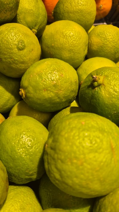 This media file is a close-up, top-down view of a pile of limes. The limes are predominantly a vibrant green, with some appearing lighter yellow-green. Their textured skin is clearly visible, and some have small blemishes or a slightly rough surface. The lighting suggests it might be indoors or under artificial light, as there are strong highlights on the fruits, creating a glossy appearance. There are no people visible in the image. The scene is an arrangement of fresh produce, likely in a market, grocery store, or a home kitchen. The background is dark and out of focus, allowing the limes to be the sole subject. The focus is sharp on the foreground and midground limes, highlighting their spherical shapes and the way they are stacked upon one another. No text is visible in the image.