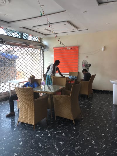 This image depicts the interior of a commercial establishment, likely a cafe or restaurant, located in Ede, Nigeria. The flooring consists of dark, possibly black, tiles with irregular white veining. The walls are light-colored, and the ceiling is white with recessed square panels and embedded lighting fixtures. A string of small, multi-colored decorative ornaments hangs across the ceiling.

On the left side, a large opening is partially secured by a raised metal roll-down security shutter, revealing an outdoor street or market area. Adjacent to this opening, a small, white rectangular table is visible. A male individual is seated at this table, facing generally right. He wears a blue short-sleeved shirt, dark trousers, dark sandals, and a light-colored cap. His hands are resting in his lap or on the table.

In the mid-ground, multiple light-brown, woven rattan-style chairs are arranged around square tables with glass tops. A female individual stands near one of these tables, leaning slightly over it. She wears a white sleeveless top and a patterned skirt. Her dark hair is styled in long braids or dreadlocks.

Further to the right, another male individual is seated in a rattan chair, facing away from the camera towards the wall. He wears a dark, patterned long-sleeved shirt and is observed looking down at a mobile phone in his hands. A standing electric fan is positioned on the floor near this individual, angled towards the right. Behind the fan, an orange horizontal blind covers a window. Electrical wiring is visible on the wall, leading to a white wall-mounted box and a smaller red square object. In the far right corner, a white pedestal sink is present.