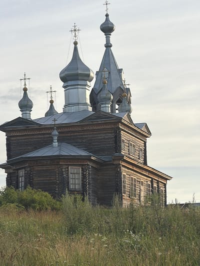 This is a vertical, eye-level, outdoor photograph of a traditional wooden Russian Orthodox church with multiple onion domes topped with crosses. The church's exterior is constructed from dark, weathered logs, and features several small, multi-paned windows. The domes are silver, and also topped with decorative crosses. The church is situated on a grassy hill with dense, dry vegetation and scattered wildflowers to the right. The sky is overcast and pale. The overall impression is one of rustic simplicity and age, likely taken in the daytime under diffused light. No people are visible in the image, and there is no visible text. The location is identified as Cherdyn, Russia.