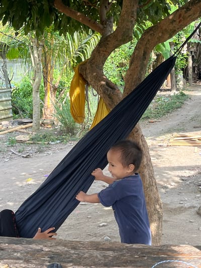 An outdoor scene in Desa Batu Layar, Indonesia, depicts a young child interacting with a hammock suspended from a tree. The child, appearing to be a toddler, wears a dark blue t-shirt and is positioned on the right, facing left, with both hands grasping the side of a black fabric hammock. The child has short, dark hair and is looking towards the hammock with a slight smile. A second individual is partially visible inside the black hammock, with their lower body and hands, wearing dark attire, resting.
The hammock is secured to the sturdy, multi-branched trunk of a large tree dominating the center-right of the frame. Another yellow fabric item, possibly a second hammock or cloth, is draped or tied around a branch of the same tree further back. The ground is a light brown dirt path or area, with scattered small stones and some sparse vegetation. In the background, lush green foliage and several trees, including a palm-like variety, are visible. To the far left, a light-colored structure, possibly a container or wall, is partially seen. A rough wooden surface, possibly a bench or table, occupies the bottom foreground. The scene is brightly lit by natural sunlight, casting distinct shadows.
