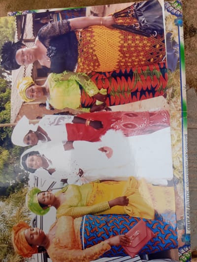 The image shows a group of women posing for a photograph, likely at an outdoor event or gathering. The women are dressed in vibrant traditional attire, with headwraps and elaborate clothing in various colors and patterns, including yellow, green, red, and blue. Some women are wearing necklaces. One woman in the foreground is holding a pink clutch bag. The background appears to be a rural or semi-rural setting, with hints of buildings constructed from corrugated iron and possibly some vegetation. The photograph itself is slightly damaged and creased, suggesting it may be an older print. A watermarked text is visible on the right side of the image.