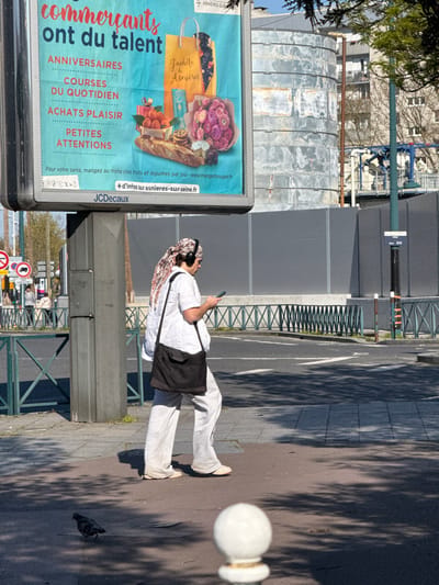 The image depicts an outdoor urban scene in Asnières-sur-Seine, France, under bright sunlight, casting distinct shadows. A large JCDecaux advertising billboard on a concrete pillar stands prominently in the left midground, displaying a light blue poster promoting local businesses in Asnières-sur-Seine, with French text and illustrations of food, flowers, and gifts. To the right of the billboard, an individual walks on a paved sidewalk, moving from left to right. This person wears a white short-sleeved top, light grey long pants, and light-colored flat shoes. A patterned white and brown headscarf is worn, with black over-ear headphones placed on top. A black messenger bag is slung over their left shoulder. The individual is holding a dark smartphone with both hands, looking down at the screen. In the immediate foreground on the pavement, a dark grey pigeon stands to the left of the walking person, and a white spherical bollard top is partially visible in the bottom right. The background includes an asphalt street, a low green metal barrier, a dark green utility pole with a 'RUE BAS' sign, a large grey industrial-style cylindrical structure, and a distant multi-story residential building with balconies.