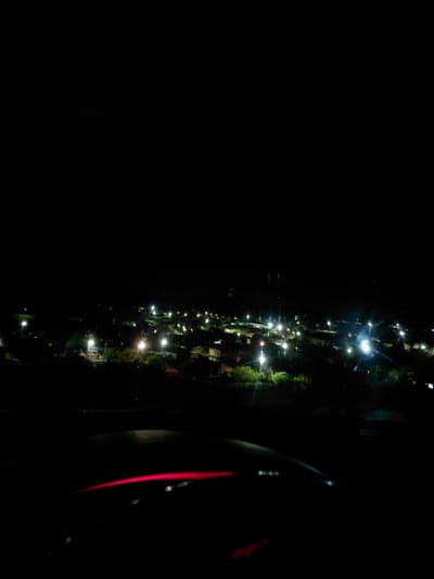 The image shows an extremely dark night scene, with a narrow strip of illuminated features running vertically along the right side of the frame. This strip appears to be a street, possibly in Dositeevo, Bulgaria, lit by scattered streetlights. The lights suggest the presence of buildings and vegetation along the street. A blurry, curved object with a red stripe is visible on the extreme right side of the frame, possibly a part of a vehicle. The left side of the frame is almost completely dark.