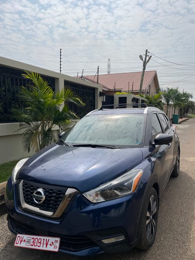 A dark blue Nissan Kicks SUV is parked on a paved residential street in Adenta Municipality, Ghana, under an overcast sky with scattered clouds. The vehicle faces forward, displaying a Ghanaian temporary license plate, "DV-3091V," affixed to the lower front bumper below the chrome-trimmed Nissan grille. The windshield and hood exhibit a light coating of dust or dirt. Silver roof rails are visible on the vehicle's roof.
To the left, a light-colored concrete perimeter wall topped with black ornate wrought iron fencing and a multi-strand electric fence system encloses a property partially obscured by green palm trees. In the middle ground, behind the wall, a building with a reddish-brown gabled roof is visible, also appearing to have security fencing. To the right, further down the street, a utility pole with overhead power lines extends upwards, and a green waste bin is situated near another property wall. A large lattice-style electricity pylon is discernible in the distant background. No individuals or active interactions are visible.