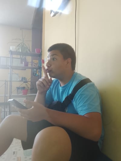 A person is sitting against a yellow wall, holding a smartphone in their left hand and bringing their right index finger to their lips in a shushing gesture. The person is wearing a light blue t-shirt and dark shorts, with a black strap over their right shoulder. In the background, a multi-tiered shelf unit contains various potted plants and other items. The scene appears to be indoors, possibly in a residential setting in Kazichene, Bulgaria, given the context. The lighting suggests it is daytime, with a bright light source from the upper left.
