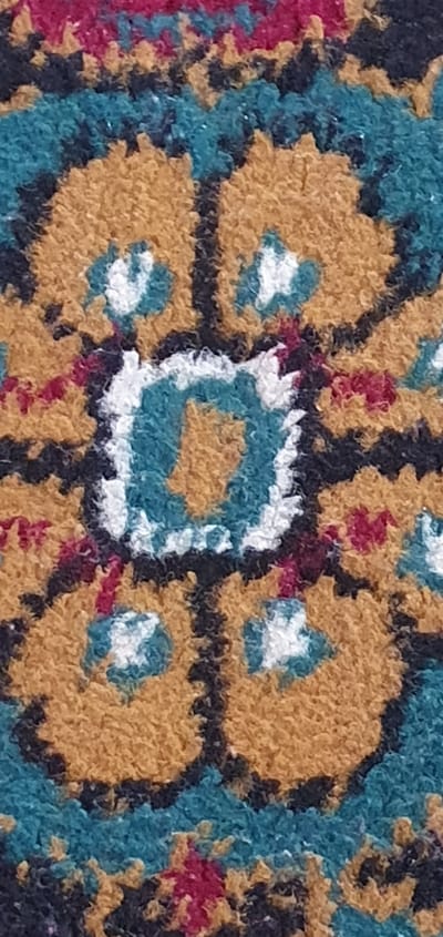 This is a close-up, high-angle shot of a woven rug with a complex geometric pattern. The rug features a central square motif with a turquoise interior, outlined in white and then a darker blue. Surrounding this central element are four large, rounded, ochre-colored shapes that appear to be stylized flowers or medallions, each with a small white dot at its center. These ochre shapes are bordered by a teal-colored pattern that creates a serrated edge. Interspersed within the design are small flecks of dark blue and maroon, adding depth and texture to the weave. The texture of the rug is visibly shaggy and tactile, suggesting it is made of thick yarn or fibers. The lighting is even, indicating an indoor setting, and there are no visible shadows that would suggest a specific time of day. The image is abstract, focusing solely on the intricate details of the textile. There is no visible text or any indication of people, animals, or a broader scene or event. The location context provided, Shurtang, Tajikistan, suggests the rug might be a traditional piece from that region.