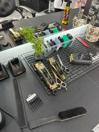 This image depicts the interior of a professional barbershop station located in Kumanovo, North Macedonia. The scene is dominated by a black, textured countertop featuring a horizontal mirror extending across its mid-section.

On the countertop, arranged primarily on a black rubber mat embossed with the "WAHL" logo, are various barber tools. In the foreground, from left to right, are two black Wahl brand charging docks, a long black fine-toothed comb, a black Wahl clipper guard labeled "2 (6mm)", and a black-handled brush with stiff black bristles.

Further back, on the "WAHL" mat, are two gold-finished Wahl brand cordless clippers/trimmers, specifically a "Magic Clip Cordless" and a "Detailer". Adjacent to these are three pairs of barber scissors, two with gold handles and one with silver handles. A black Wahl "Vanish" shaver is also present. To the left of the clippers, within a clear plastic organizer, are multiple colored clipper guards, including black, grey, light blue, and green. A small artificial plant in a dark pot sits on the mat, reflecting in the mirror.

On the right side of the mat, a red-handled straight razor and a small black folding utility knife or object are visible.

Reflected in the mirror, in the background, are a black, button-tufted barber chair, the upper torso of a person wearing a white short-sleeved shirt, a gold-colored spray bottle featuring red and blue barber pole stripes, and two skull-themed tool holders with steampunk-style gear embellishments, each containing multiple silver scissors and other small implements. The reflection also shows additional "WAHL" mats and tools.
