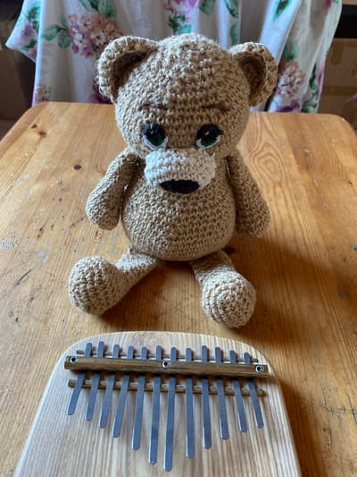A handmade crocheted bear sits on a wooden table in front of a kalimba. The bear is light brown, with green eyes and a white muzzle. It is facing the camera, with its arms resting on its knees and its legs splayed out. The kalimba is made of light-colored wood with metal tines. Behind the bear and table, a floral patterned fabric is visible. The lighting suggests an indoor setting with natural light. The scene is set in the city of Zelenodol'sk, Russia.
