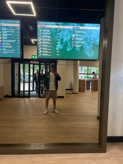 The image, rotated 90 degrees counter-clockwise, captures an indoor scene, likely a cannabis coffeeshop or dispensary, characterized by digital menus and reflections in a glass partition. The space features a light-colored wooden floor and bright overhead lighting.
Two prominent digital screens display detailed menus. The left screen lists "HASJ" and "VAPE PEN" options, including strains like "BLUE ZUSHI X MAC: THC 50.05%" and "PIATTELLA THC 62.05%," with prices per gram. The right screen presents "JOINTS" and "EDIBLES." Joint selections such as "THE HULK JOINT" and "PARTY JOINT HAZE" are priced from €3.00 to €7.50, with discounts for bulk purchases. Edible items include "CHOCOLATE BAR (Very Strong - Flavour: Milk)," "SPACE CAKE (Flavour: Red Velvet)," "STROOPWAFEL," "HAPPY TEDDY GUMMY," and "DOPE BROWNIE," ranging from €6 to €31.50. Visible text also includes rules like "Minimale besteding" (minimum spend) and "Consumptie verplicht" (consumption mandatory). A green exit sign is visible between the screens.
A young man is prominently reflected in the glass partition, facing the menus. He wears a dark cap, a light-colored t-shirt with a "18" graphic, a dark jacket, light shorts, and white sneakers. He holds a red-cased smartphone, possibly taking a photograph or reviewing the menu. Beyond the glass, an outdoor scene with green trees and a street with parked cars indicates it is daytime. Faint reflections of indoor seating and other individuals are also discernable. The setting in Ede, Netherlands, aligns with the legal operation of such an establishment.