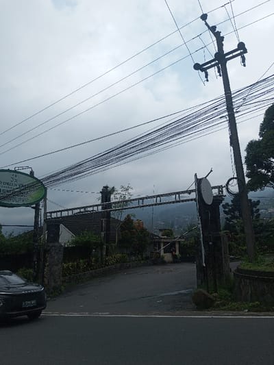 An overcast day illuminates the entrance to "Green Village." A car pauses on the asphalt road, its license plate reading "S 1769 BAS." The entrance features stone pillars and a dark wooden structure. A guard booth is visible in the distance. Overhead, a dense web of power lines stretches across the sky, connected to a utility pole. The background reveals a sloping landscape with trees.