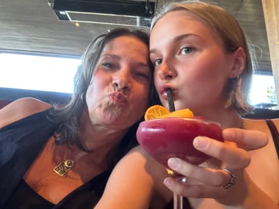 Two women pose closely together, with a cocktail glass held near the younger woman's face. The cocktail is a deep red color and garnished with an orange slice. The younger woman is drinking from the cocktail using a straw. The older woman has her lips pursed and seems to be trying to kiss the younger woman. The younger woman has fair skin, blonde hair, and a nose piercing. The older woman has darker hair, and wears a gold necklace. The photo is taken from an upside-down angle. Based on the architectural details visible in the background, the location may be an outdoor patio space. The location in Kelowna, Canada is not evident from the image.