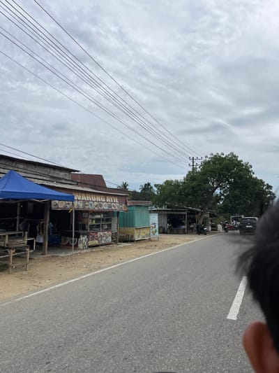 The image shows a roadside scene in Tanahterban, Indonesia, on a cloudy day. In the foreground, the right side of a person's head and shoulder are visible, suggesting the photo was taken from a moving vehicle. A paved road with a white line runs through the center of the frame. To the left of the road are several small roadside businesses. The closest business, covered by a blue tarp, appears to be a seating area with wooden tables and benches. Next to it is a shop with a sign that reads "WARUNG AYIE," displaying an array of food pictures. Further down the road, more stalls and shops are visible, including one with a "BESTTEA" sign. A large tree dominates the background on the right side of the road, with other greenery and buildings in the distance. Several motorcycles and a black SUV are parked or moving along the road, indicating a moderately active area. Power lines stretch across the sky above the scene.