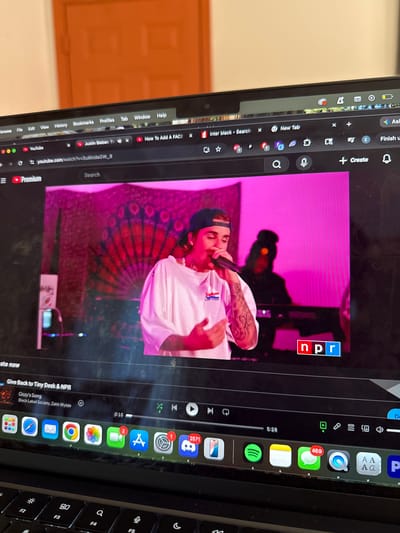 A laptop screen displays a YouTube video of a musical performance by Justin Bieber, featuring an NPR logo, set against a pink backdrop with a patterned design resembling a peacock feather fan. The performer, wearing a light-colored shirt and a backward cap, holds a microphone. The video shows timestamps "0:15" and "5:28", with the title "Give Back to Tiny Desk & NPR" and "Ozzy's Song" by "Black Label Society, Zakk Wylde" visible below. The laptop screen appears smudged or dirty.
On the left side of the screen, various browser tabs are open, including "inter black - Search," "New Tab," "How To Add A FACE," and "Justin Bieber: Tir". The YouTube URL "youtube.com/watch?v=Su6kidaGW_8" is visible. A YouTube Premium banner is also present. The browser's menu bar shows "Chrome," "File," "Edit," "View," "History," "Bookmarks," "Profiles," "Tab," "Window," and "Help."
The laptop's dock or taskbar is visible on the right, displaying numerous application icons. Several icons show notification counts: Mail (1), Messages (469), Settings (1), App Store (2), and Discord (2571). Other visible icons include Safari, Chrome, Photos, Spotify, Notes, and a generic app icon. The keyboard is partially visible at the bottom right, showing keys like F2, F3, F4, and F5.
The background shows a light-colored wall or door, indicating an indoor setting. The image is taken at an angle, showing the laptop screen clearly, along with its surrounding parts.
