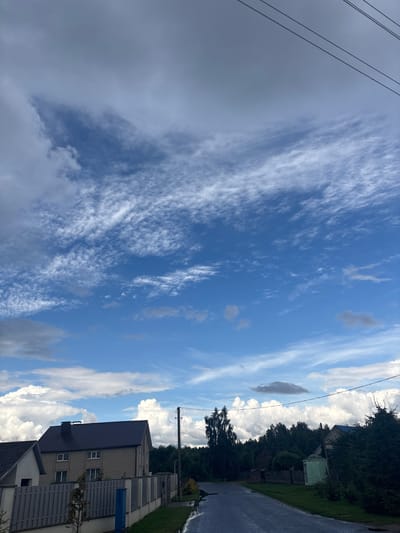 The image depicts a street scene in the Kapylsky Sel'skiy Sovet, Belarus. The sky is partly cloudy with a mix of large cumulus clouds and wispy cirrus clouds. Power lines are visible in the upper right quadrant of the image. A two-story brick house with a dark roof and a fence in front of it is located on the left side of the street. To the right of the house, a paved road stretches into the distance, flanked by trees and other residential buildings. The road appears wet, suggesting recent rain. The overall impression is of a rural or suburban residential area under a dynamic sky.