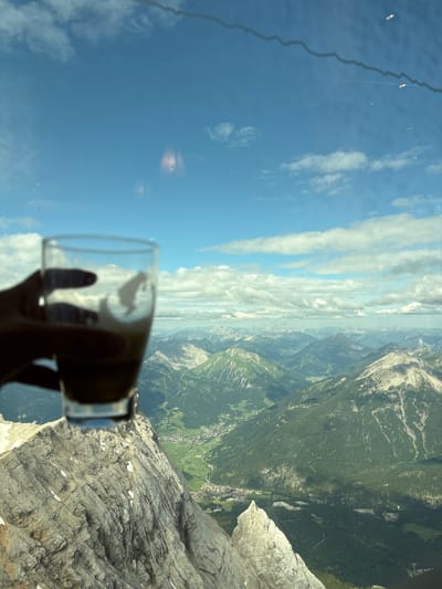A hand holds a glass of coffee in the foreground, with a blurred mountain landscape and a blue sky with white clouds in the background. The mountain landscape appears to be the Alps, with lush green valleys and snow-capped peaks. The city of Ehrwald, Austria, is visible in the distance, nestled in a valley. The image seems to be taken from a high vantage point, possibly a cable car or a mountain peak. The overall mood of the image is one of relaxation and enjoyment of nature's beauty.