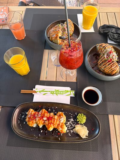An overhead, eye-level photograph depicts a dining table laden with food and drinks, presumed to be in Basel, Switzerland. The table is constructed from light wooden slats and covered with dark grey, textured placemats. In the upper background, the lower torso and legs of a seated individual, wearing dark shorts and a white shirt with a blue/black pattern, are partially visible.
Four drinks are present: two clear glasses containing bright orange juice, one clear glass with a light pink beverage and ice, and a stemmed glass holding a vibrant red cocktail garnished with dried petals and a black straw.
Food items include two dark grey bowls, each containing a large, deep-fried roll, likely sushi, wrapped in dark seaweed, drizzled with a light brown sauce, and heavily topped with green and red microgreens. In the foreground, an oval black plate features six sushi rolls, coated in an orange sauce, sprinkled with red and white flakes, and accented with dark sauce drizzles. Accompanying condiments consist of a small white dish of dark soy sauce, a dollop of green wasabi paste, and a pile of pale pickled ginger. A pair of wooden chopsticks rests on a white paper sleeve bearing a green bamboo graphic and the Japanese text '天然の竹' (natural bamboo). A crumpled black plastic object lies on the table's upper right side.
