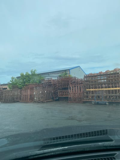 The image shows an outdoor storage area filled with metal racks and wooden pallets. The racks are of various sizes and appear to be made of rusted metal. Many of them are stacked high, creating a cluttered but organized appearance. The ground is wet and reflective, suggesting recent rain. In the background, there is a building with a grey facade and a blue roof, possibly an industrial or commercial structure. Trees with green foliage are visible behind some of the racks, and a blue cart with wheels is positioned near the stacked racks on the right side of the image. The sky is overcast and a pale blue with some clouds. The foreground of the image shows the dashboard of a vehicle, indicating the photo was taken from inside a car. There are no people visible in the image. The scene appears to be in an industrial or storage yard within the city of Shchyolkovo, Russia.