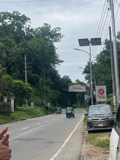 The image depicts a wide, paved road in Sylhet, Bangladesh, flanked by lush green trees and foliage on either side. The road has white lane markings. In the foreground on the left, a person's hands are visible, with a thumb and index finger extended. On the road, three green auto-rickshaws are in motion, with two further in the distance and one closer to the center. A dark-colored van is parked on the right side of the road. Overhead, electrical wires are strung between poles, and two solar panels are mounted on a pole. Various signs are visible along the roadside, including a circular speed limit sign indicating '40' and other signs with text in Bengali. A large banner with text that appears to read "Leading University" is also visible in the distance. The sky is overcast.