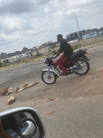 A man stands next to a red motorcycle on a paved road in the Aso Housing Estate, Nigeria. The man is wearing a black shirt and red pants. He appears to be working on the motorcycle. Behind the man and the motorcycle are rows of houses. The sky is cloudy. A vehicle mirror is visible in the lower right corner of the image. Debris is also visible next to the vehicle mirror.