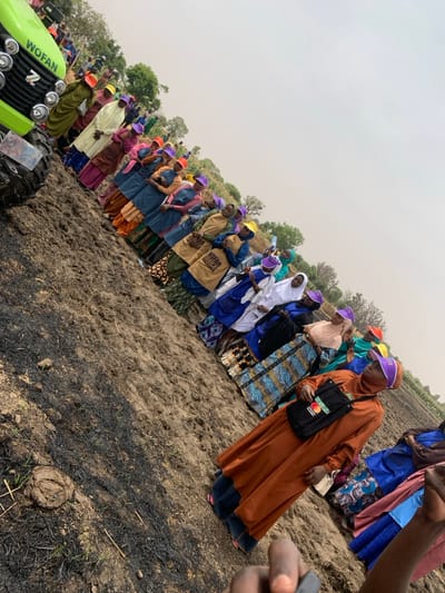 A group of women are gathered on a field in Ringimawa, Nigeria. They are standing in a line, facing the camera. They are wearing colorful clothing. Some of them are holding yellow bags with the words "WOMAN IN AGRICULTURE" printed on them. There is a green tractor in the background. The field is brown and dry, and there are some charred patches of vegetation. The women are listening attentively as someone speaks. The sky is overcast. The coordinates are: Latitude: 11.666, Longitude: 8.414. This appears to be an agricultural training event or a demonstration. The presence of the tractor and the phrase "WOMAN IN AGRICULTURE" on the bags suggests an initiative focused on empowering women in agriculture.