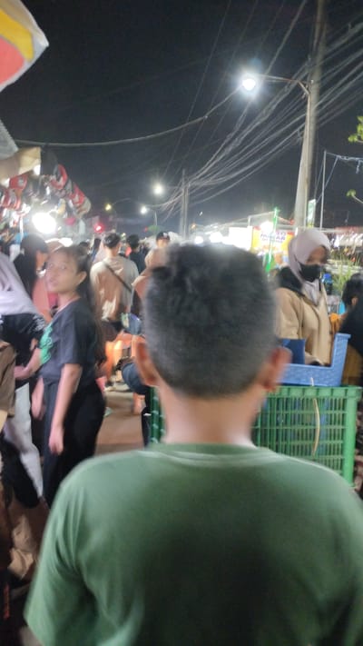 The image depicts a crowded outdoor scene at night, likely a market or festival in Sukodono, Indonesia. The foreground is dominated by the back of a person wearing a green t-shirt, with their head and shoulders out of focus.
Behind them, a diverse group of people are visible, milling about under artificial lighting. A young woman with long, dark hair, wearing a dark t-shirt and dark pants, is looking towards the camera with a curious expression. She is surrounded by other individuals, some blurred by motion or distance. Further in the background, a woman wearing a light-colored hijab and a black mask is visible.
The setting appears to be a bustling public area with stalls or vendors. Several streetlights illuminate the scene, casting a warm glow. Overhead, a network of electrical wires is prominent, strung between poles, which is common in many Indonesian urban or semi-urban areas. One stall displays items that are difficult to discern due to the lighting and distance, but appear to be hanging products.
A green plastic crate is visible in the mid-ground, suggesting goods or supplies are being organized or transported. The overall atmosphere is lively and somewhat chaotic, characteristic of a busy night market or event. The time of day is clearly night, indicated by the dark sky and the reliance on artificial lights. There are no overt indications of the weather, but the lack of rain suggests it is likely dry.
Visible text includes "PESO MILAR OF KEDIRI" on a sign in the background, providing a specific geographical clue to the location. The mood of the people is difficult to ascertain with certainty due to the image quality and the focus, but the general impression is one of activity and perhaps mild curiosity.
