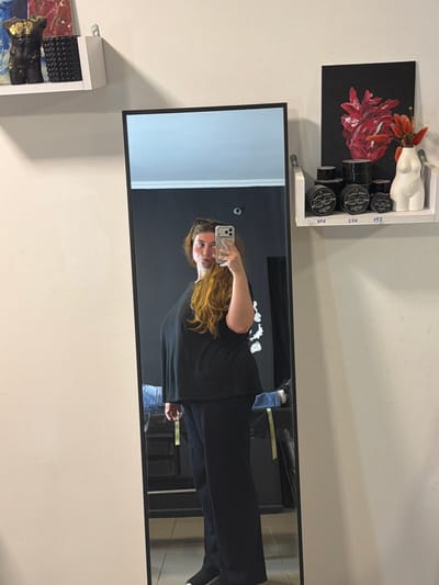 This media file shows a person taking a selfie in a mirror. The person has long, reddish-brown hair and is wearing a black long-sleeved shirt and blue jeans. They are holding a white smartphone in their right hand, angled to capture their reflection. The background of the mirror is dark and appears to be a studio or retail space.
On a shelf above and to the left of the mirror, there are several small black jars with white labels, arranged next to a white, torso-shaped object adorned with dried red flowers. A black canvas with a red floral painting is also visible on the shelf.
To the left of the mirror, a white shelf holds what appears to be abstract art, featuring darker tones with metallic gold accents. Visible text on the jars on the upper shelf reads "TATTOO CREAM" and some numbers, possibly prices, such as "150", "250", and "350". The overall setting suggests an art studio, tattoo parlor, or a retail space that sells art and related products. The lighting is bright, suggesting daytime. The mood appears casual and everyday.