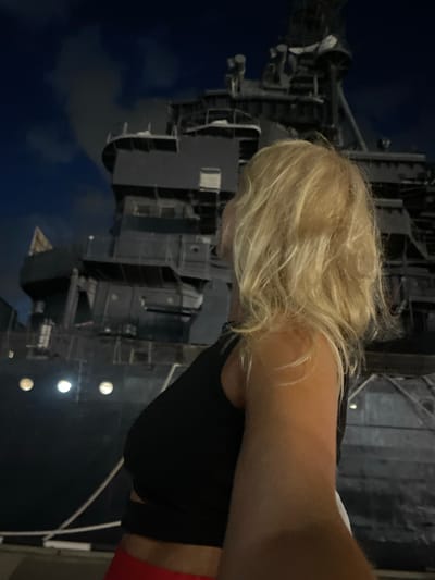 A woman with blonde hair is standing in front of a large, dark ship at night. The ship appears to be a warship or a large vessel with multiple levels and structures. The woman is facing away from the camera, looking towards the ship. She is wearing a black tank top and red bottoms. The environment is dark, with some lights on the ship visible. The sky above is dark with some clouds. The location is not Verkhnyaya Stavropol'ka, Russia.