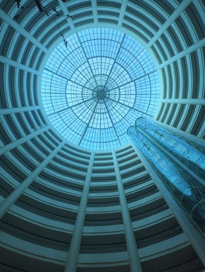 This is a low-angle, wide shot looking up at the interior of a large, modern building with a circular atrium. The ceiling is a large, domed glass structure with a grid pattern, allowing natural light to filter in. The walls of the atrium are tiered in a circular fashion, resembling balconies or walkways, with white cylindrical columns supporting the structure. Two glass elevator shafts, reflecting the light and surrounding architecture, rise through the center of the atrium. The overall color palette is dominated by cool blues and grays, suggesting artificial lighting and the color of the glass. The scene is devoid of people, emphasizing the architectural elements. The image is likely taken within the Dali City China International Exhibition and Convention Center.