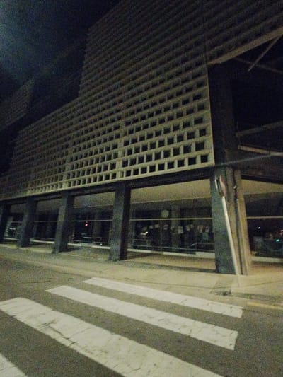 The image captures a building at night with a distinctive perforated facade, likely a Brutalist architectural style. The facade features a grid of square openings creating a repetitive pattern. The building is elevated on pillars, with a ground-level storefront behind glass. A crosswalk in the foreground suggests an urban setting. The scene's location is likely Rubí, Spain. No individuals are clearly visible, though there may be people inside the building. The lighting is dim, emphasizing the architectural features.