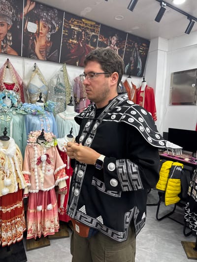 An adult male stands in profile, facing left, inside a costume rental or photography studio in Fenghuang Ancient Town, China. He is wearing a black tunic-style garment with intricate silver geometric patterns and circular silver disc embellishments on the chest, sleeves, and shoulder flaps. He has short, dark, curly hair, glasses, and a serious expression, with his hands clasped in front of him. A white smartwatch is visible on his left wrist, and olive-green shorts along with a two-tone fanny pack are visible beneath the tunic.

The background features a retail environment with several mannequins displaying traditional Chinese costumes. To the left, child-sized costumes include a pink fur-trimmed coat with a large red bow and a floral headpiece, and a reddish-orange long dress. Adult female costumes in light blue and green with delicate fabrics and floral details are also visible. Above the costumes, a row of five large framed photographs on the wall depicts models in elaborate traditional attire, including ornate headpieces. To the right, a yellow quilted jacket hangs on a black stand, and a metal box is mounted on the white wall. The ceiling features black track lighting, and the floor is light-colored tile.