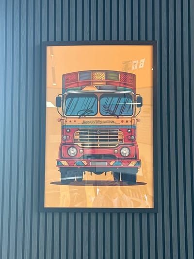 A vertical photograph captures a framed, colorful artwork depicting a stylized truck, hanging on a dark grey, horizontally grooved wall. The artwork inside the frame is rotated 90 degrees clockwise, so the front of the truck faces the left side of the image. The truck is vibrantly colored with a red cab, orange body sections, blue windshields, and multi-colored decorative patterns in yellow, green, and black. It features a prominent grille, circular headlights, and visible windshield wipers.

Significant reflections on the picture's glass obscure some details. A bright, linear glare runs horizontally across the lower third of the framed image, and a distinct small white reflection is visible near the top center of the frame. Faint, stylized text resembling "BLG" or similar characters is visible on the upper left side of the truck's body within the illustration. Small, decorative script is also present above the windshield, but it is illegible.

The wall on which the picture hangs features a consistent dark grey or blue-grey tone with noticeable horizontal ridges or slats, suggesting a modern or textured finish. The overall setting is indoors, with ambient light causing the reflections on the frame. The location context is Pitt Meadows, Canada. No activity is depicted beyond the display of the artwork.