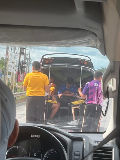 The image shows a side-view of a tuk-tuk filled with several young people, likely students, as indicated by the text "THASALAPRASITSUKSA SCHO" on a purple shirt. The tuk-tuk is dark-colored with yellow and black upholstery. One person in the foreground wears a bright yellow t-shirt and black shorts, facing away from the camera. Other individuals are visible inside the tuk-tuk, some wearing blue shirts. The scene appears to be a street in Tha Sala, Thailand, with traffic lights and road signs visible in the background. The sky is overcast, suggesting a cloudy day. The dashboard of a car, presumably the one from which the photo is taken, is visible on the right side of the frame, showing a speedometer with a speed of 0 km/h, the time 4:12, and the number 319. The overall impression is of a mundane moment of transportation, possibly during a school day.