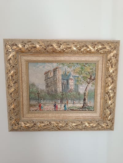 The image shows a framed painting of Notre Dame Cathedral in Paris. The painting depicts the cathedral's facade with its iconic towers and gothic architecture. In the foreground, there is a tree and a paved area where several figures are walking. The figures are small and stylized, suggesting a bustling urban scene. The painting style appears to be impressionistic, with visible brushstrokes and a vibrant color palette. The frame of the painting is ornate and golden, with intricate carvings of leaves and vines. The painting is hung on a plain white wall, with the light casting a shadow on the wall below. The location in the city of Vitoria-Gasteiz, Spain is not depicted in this image; the painting itself portrays a scene in Paris, France.