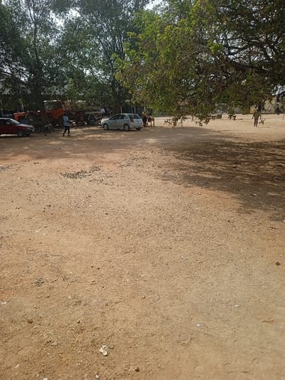 The image displays an expansive outdoor area in Mubi, Nigeria, characterized by an unpaved reddish-brown dirt ground strewn with small stones and debris. Overhead, a bright, clear sky is visible. Numerous large trees with green foliage frame the scene, particularly on the left and right, casting significant shadows across the ground on the right side.
In the left mid-ground, a red sedan is partially visible, parked beneath a tree. Behind it, a prominent older, orange-colored utility vehicle, consistent with a truck or tractor, is parked. Another smaller, darker vehicle is partially obscured behind the orange utility vehicle. A person wearing a blue shirt and dark trousers is walking from left to right in front of the orange utility vehicle. Several other individuals are observed standing or sitting in the vicinity of these parked vehicles in the background, including one person in a white top.
In the mid-ground, a light-colored, possibly silver or light gray, hatchback car is parked, facing right. To the right of this vehicle, a small group of individuals is gathered around a low, dark structure, possibly a table or stand. In the far right background, a lone individual is walking from right to left across the open dirt area, accompanied by a small, dark animal, consistent with a dog. The scene collectively suggests an informal outdoor gathering or parking space.