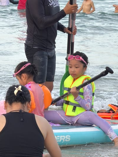 In Dong Ninh Hoa, Vietnam, on a body of water, a young girl in a bright yellow life vest and purple leggings sits on a blue paddleboard. She wears pink goggles and holds a paddle with a black handle. Beside her, another child in a pink shirt and orange flotation device is partially visible. In the background, an adult wearing a black long-sleeved shirt and dark shorts stands in the water, holding two poles. Another young person, shirtless, stands further in the water. The environment appears to be a beach or coastal area with gentle waves.
