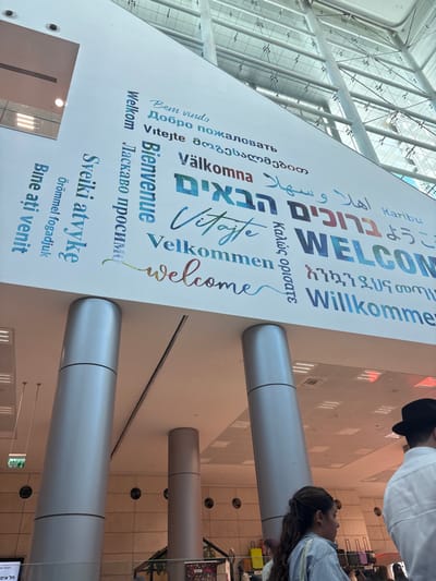 The image depicts the interior of a modern public building, consistent with a major transportation hub or convention center such as Jerusalem's Yitzhak Navon Railway Station, located in West Jerusalem, Israel.

A large, white, rectangular sign prominently displays the word "WELCOME" in approximately 18 different languages, including Hebrew (ברוכים הבאים), Arabic (أهلاً وسهلاً), English, Russian (Добро пожаловать), French (Bienvenue), German (Willkommen), Swedish (Välkomna), Portuguese (Bem vindo), Amharic, and Japanese (ようこそ). This sign is affixed to a high, white ceiling which incorporates recessed lighting and a skylight-like structure composed of metal beams and glass panels, allowing natural light into the space. Several large, silver-gray, cylindrical pillars, tapering slightly, rise from the floor to support the overhead structure. A green "EXIT" sign, featuring Hebrew text (יציאה), is visible on a wall section to the far left.

In the lower right foreground, the back of a male individual wearing a white shirt and a black fedora hat is visible. Immediately to his left, a female individual with long, dark, curly hair tied back is observed in profile, facing right, dressed in a light-colored striped shirt. Other indistinct figures are partially visible in the background among what appear to be various objects, possibly luggage or commercial displays. The overall scene is brightly lit and indicates a spacious, contemporary public area designed for movement and transit.