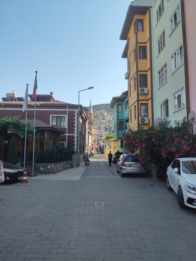 This is an outdoor, eye-level, vertical shot of a narrow street in Osmangazi, Turkey. The street is paved with gray interlocking stones. On the right side of the street, several buildings of varying heights and colors are visible, painted in shades of green, yellow, and pink. Some buildings have balconies and air conditioning units. Lush pink flowering bushes are in the foreground on the right, obscuring the lower portion of the buildings. A white car is parked on the right, and a silver car is parked further down the street.

On the left side of the street, there is a darker colored building with a wooden porch and a sign that reads "CUMALIKIZIKLI KÖY KAHVALTISI". Two flags are mounted on poles in front of this building. Further down the street on the left, more buildings and a distant view of a hill covered in houses are visible. A minaret of a mosque is also visible in the distance.

Several people are present in the scene. In the middle ground, a woman is pushing a small wheeled cart. Two young men are walking down the street further in the distance. A black car is parked on the left side of the street.

The sky is clear and blue, suggesting it is daytime with good weather. The overall mood is peaceful and suggests a quiet, everyday moment in a town. The time of day appears to be late afternoon or early evening, given the lighting. The license plate of the silver car reads "16 GL 690". The license plate of the black car reads "34 NVY 13". A sign on the water tank on the left reads "346 90 06".