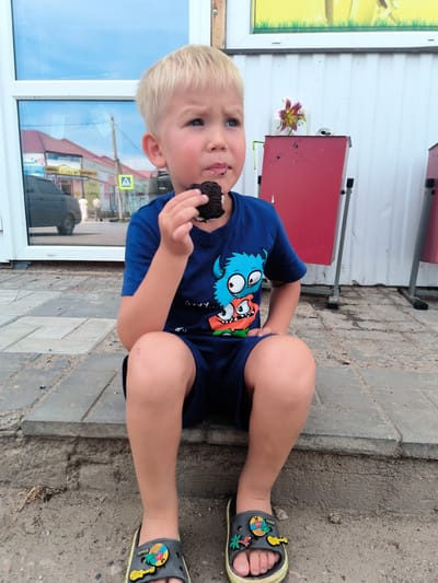 A young boy with blonde hair is sitting on a concrete step outdoors, eating a chocolate cookie. He is wearing a dark blue t-shirt with cartoon monsters and dark blue shorts, along with dark gray Crocs sandals. The background shows a building with a large window reflecting a street scene with buildings and a yellow traffic sign, suggesting the location is in the city of Arkhangelskoye, Russia. There is also a red metal structure with a flower next to it. The ground is made of concrete pavers and dirt.