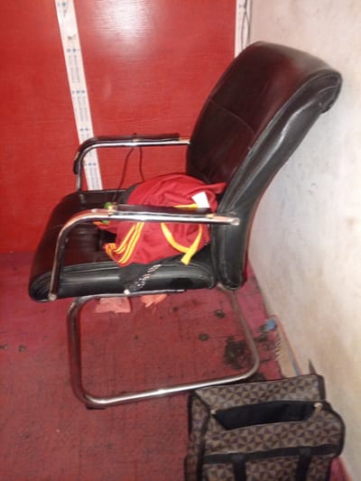The image depicts a disheveled interior scene. A black leather office chair with chrome accents is positioned on a red carpet. A red shirt with yellow stripes is draped over the back and seat of the chair. To the right of the chair, a patterned tote bag with black straps sits on the floor. A portion of a red wall is visible in the background, with a white and blue striped tape measure affixed to it. The scene appears to be located in Lankoviri, Nigeria.