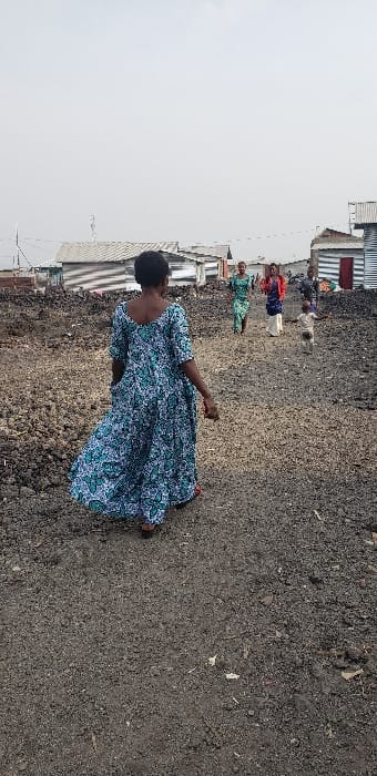 The image depicts an outdoor scene in Buzove, Democratic Republic of the Congo. A wide dirt path, composed of dark earth and lighter gravel, extends from the foreground into the midground. The ground surrounding the path is dark, loose, and uneven. In the background, numerous single-story structures constructed from corrugated metal sheets serve as informal housing; some structures feature red or dark-colored door sections. The sky is uniformly light grey, indicative of overcast conditions.

In the immediate foreground, a person with dark skin and short, dark hair is positioned with their back to the viewer, walking forward on the dirt path. This individual wears a loose, floor-length dress patterned in teal and white, with dark red footwear partially visible.

In the midground, five additional individuals are visible on the same path, walking towards the foreground. These include a person in a green patterned dress, another in a dark blue top with a light skirt and a red overlay, a small child in light-colored clothing walking ahead of the adults, and two further individuals to the right. All individuals are dressed in everyday attire. No direct interaction between the foreground and midground groups is depicted; all are moving within the shared open space.