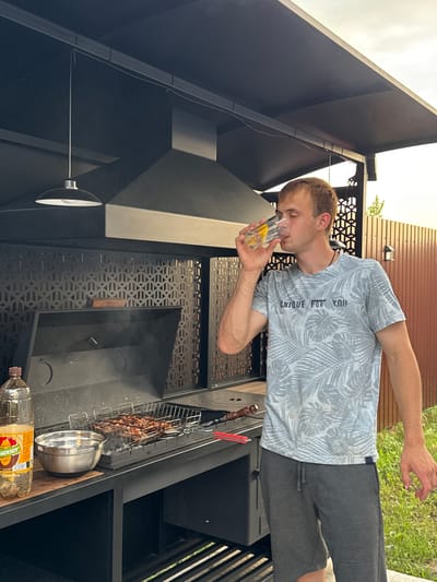 In Starokorsunskaya, Russia, a young man with blonde hair stands beside a black outdoor grill. He is wearing a light blue and white patterned t-shirt and dark gray shorts, and is drinking from a clear glass containing a yellow liquid and ice. The grill is smoking, with meat cooking on a metal rack. To the left of the grill, there is a large plastic bottle of a yellow beverage and a stainless steel bowl. The scene is outdoors, with a wooden fence visible in the background and green grass underfoot. A black metal awning with a hanging light fixture is positioned above the grill.