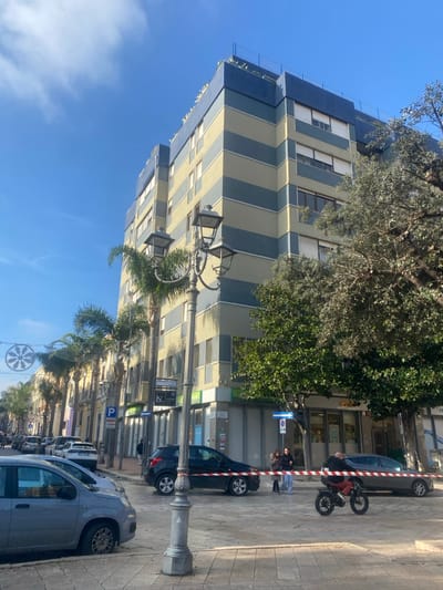This image depicts an urban street scene in Brindisi, Italy, under a clear blue sky with scattered white clouds. The main background feature is a multi-story building, approximately seven to eight floors high, characterized by alternating horizontal stripes of light beige and blue-grey. Its ground floor houses commercial establishments with visible storefronts.
In the immediate foreground, a grey, ornate street light post with three lamps stands on the patterned sidewalk. To the left of the lamppost, a grey hatchback is parked on the street. Further down the street on the left, a row of palm trees is visible, along with additional parked vehicles and a white, snowflake-shaped decorative light fixture suspended above the street.
The mid-ground features a section of the street cordoned off by red and white striped barrier tape. Behind this tape, a black SUV is parked to the left, and a grey sedan is visible further to the right. Two individuals are standing behind the tape, observing the scene; one wears a light blue top and dark pants, the other a dark top and dark pants. In front of the barrier tape, a male individual, wearing a dark jacket and reddish pants, is riding a dark-colored motorized bicycle from right to left across the frame. On the right side of the scene, dense green foliage from several trees partially frames the street and building. Various street signs, including a 'P' for parking and a blue one-way arrow, are present.