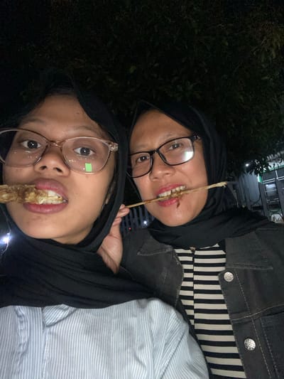 Two women are shown in a selfie. The woman on the left is wearing glasses and a black hijab, and is holding a skewer with food to her mouth. The woman on the right is also wearing glasses and a black hijab, and is holding a skewer with food to her mouth. Both women appear to be in an outdoor setting at night, possibly a street food stall in Tanggul, Indonesia. The background shows dark foliage and some blurry lights, suggesting it is nighttime. One woman is wearing a light blue and white striped shirt, and the other is wearing a black and white striped shirt under a dark jacket. The image is taken from a low angle.