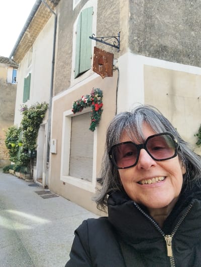 A woman with gray hair and large, square sunglasses smiles into the camera. She wears a black jacket zipped up to the neck. Behind her, a narrow European street features old stone buildings. A closed shuttered window is decorated with a Christmas garland. A rusty metal sign hangs above it. Green shutters are visible on other windows. Ivy climbs the side of one building. The street is paved with light-colored stones.