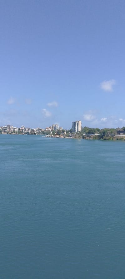 The image displays an expansive coastal cityscape across a body of blue-green water under a clear blue sky, identified as Junda ward, Kenya. The foreground is occupied by tranquil, textured water with minimal surface ripples, extending upwards from the bottom of the frame. The midground reveals a distant, developed coastline with numerous multi-story buildings, predominantly light-toned, ranging in height and architectural style. Several prominent high-rise structures are visible, particularly in the left-center. Along the immediate shoreline, small vessels or boats are discernible, moored near what appear to be docks or waterfront structures. Green vegetation is interspersed with the urban development and natural landforms along the coast. The upper portion of the image is dominated by a bright blue sky, featuring a few scattered, small white clouds. No individuals or specific active interactions are visible. The scene is illuminated by daylight.