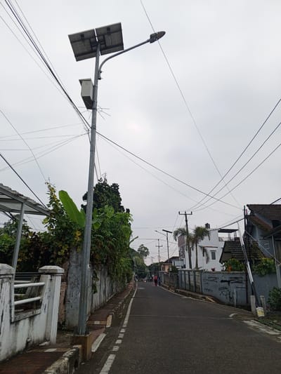 The image depicts a street scene in Pelwehera, Sri Lanka. A grey asphalt road stretches into the distance, lined with a concrete wall and sidewalk. A prominent lamp post with solar panels stands on the left side of the road, accompanied by a small grey utility box. Overhead, a complex network of electrical wires crisscrosses the sky, obscuring a cloudy atmosphere. Adjacent to the wall, lush green vegetation, including large banana leaves, thrives. In the background, residential buildings with tiled roofs are visible, interspersed with palm trees. A few people can be seen walking on the road in the distance. The overall impression is of a residential street on an overcast day.