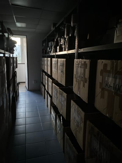 The image shows a dimly lit warehouse or storage area. The perspective is unusual, as if the camera is looking upwards at shelves laden with stacked cardboard boxes. Many of the boxes are wrapped in plastic, and some have visible labels. One box clearly shows the text "96-100". Another box has "95-96" visible.
In the foreground, at the bottom of the image, there are more boxes stacked on shelves. To the left, there is a doorway or opening leading to an area with natural light, possibly an exterior view, though it's indistinct. Bags and other items are visible in this lighter area.
The walls of the warehouse appear to be tiled or paneled in a dark, possibly metallic material. The overall impression is one of a functional, somewhat cluttered storage space, possibly during daylight hours given the light from the doorway, but the interior is largely in shadow. There are no people visible in the image.