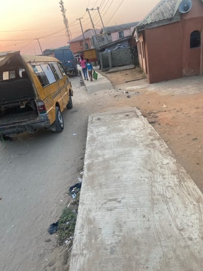 This image captures a vibrant street scene in Ojo, Nigeria, viewed from a slightly elevated perspective. The setting appears to be an urban or semi-urban area characterized by a mix of residential and commercial structures.
In the foreground, a large, rough concrete path or barrier runs diagonally across the right side of the frame, separating a dirt/unpaved road from what might be a drainage ditch or an unkempt area with sparse vegetation and visible litter, including plastic bags.
On the unpaved ground to the left of the concrete structure, a weathered yellow van, likely a "danfo" public transport vehicle common in Nigeria, is prominently parked or stopped. The rear of the van is visible, displaying faded text or numbers, possibly including "88 NIGERIA" near the taillights. The van's rear window is open, and its body shows signs of wear.
Further down the street, towards the midground, several individuals are gathered. Among them are a few children, one in a pink shirt and another in a blue shirt, standing near a dark blue van or bus. Buildings of varying heights and architectural styles line both sides of the street, some with exposed concrete blocks and others painted in earthy tones. On the far left, utility poles laden with numerous overhead power lines extend into the distance.
A loudspeaker is mounted on the upper portion of a brown-painted building on the far left side. The ground throughout the scene is predominantly unpaved and dusty. The lighting suggests either late afternoon or early evening, with a warm, low-angled sunlight casting a soft glow across the scene, particularly on the left horizon. The overall impression is that of a lively community thoroughfare.