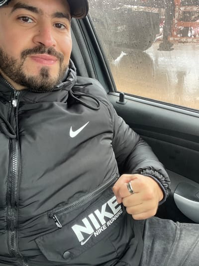 A male individual is depicted seated in the passenger seat of a vehicle in Oued Jdida, Morocco, during what appears to be rainy weather. He has short dark hair, a trimmed beard, and a mustache, and is looking directly at the camera with a slight smile. He wears a black puffer jacket featuring a white Nike swoosh logo on the chest and "NIKE NIKE RUNNING" text on a lower zippered pocket. A dark baseball cap is partially visible on his head. On his left ring finger, he wears a silver-colored ring with dark concentric detailing. The vehicle's dark interior door panel is visible to his left, with a black door lock pin positioned near the window. Through the window, the exterior environment is blurry and obscured by rain droplets, indicating wet conditions. Vague shapes in the background suggest an outdoor setting, possibly an urban or semi-urban area.