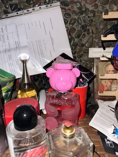 The image shows a collection of various objects, primarily perfume bottles and a document, arranged on a textured surface that appears to be stone or pebbles. In the background, there's a woven mat-like material.
The most prominent objects are several perfume bottles. One is a bright pink, teddy bear-shaped bottle, and next to it is a clear glass bottle with a red-tinted liquid and the word "MOSCHINO" visible on its label. Another clear glass bottle with a black cap is also visible, and to its right, another perfume bottle with a silver-toned atomizer. A partially visible gold-colored perfume bottle with a distinctive silver stem and clear cap is also present.
A white document, possibly a form or report, is partially visible. Text on the document includes headings such as "АКТ № 246", "ОКАЗАННЫХ ПЛАТНЫХ УСЛУГАХ", "Наименование показателя", "Дата составления", "Исполнитель", "Заказчик", and "Адрес объекта". There are handwritten entries on the document. Another smaller document or label with Cyrillic text and a barcode is also present.
In the upper left and center background, there are wooden decorative elements, one of which appears to be a cutout with text. One cutout shows a stylized depiction of a person's face and religious imagery.
The scene appears to be an indoor setting, with artificial lighting, as evidenced by the reflections on the glass bottles. No specific time of day or weather conditions can be determined. The overall impression is one of a personal collection or perhaps a disordered arrangement of items. There are no people visible in the image. The location context is Bishkek, Kyrgyzstan.